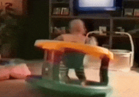 Video gif. In a living room, a baby spins wildly around in a sit-and-stand plaything. 