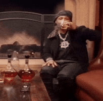 Celebrity gif. In front of a fireplace, Katt Williams sits in a leather armchair with his hand rested upon his knee and casually takes a sip of an alcoholic drink. 