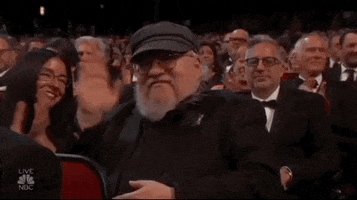 Celebrity gif. George R.R. Martin sitting in the audience at the Emmy Awards waving at us.