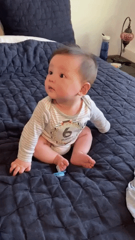 Video gif. A happy baby sitting on a bed leans back and falls, giggling.