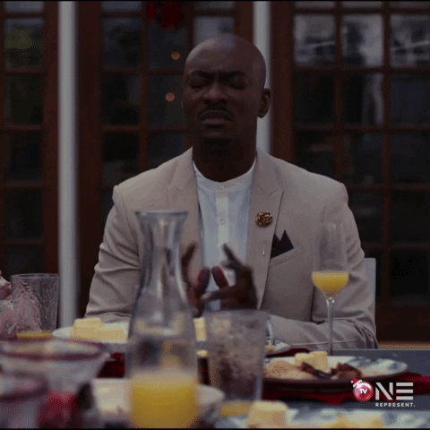 Movie gif. Christopher B Duncan as Howard in "Christmas Dilemma" sits at a brunch table with his eyes shut, holding up his hands as if to quiet people and putting his hands together in prayer.