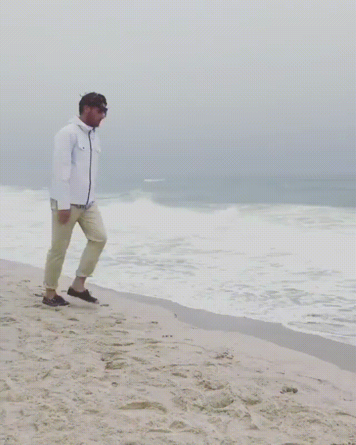 Video gif. A man stands at the edge of a sand bank, with the tide in the background. He stares at us before jumping onto the sandy ledge, letting it slowly carry him down and out of sight. 