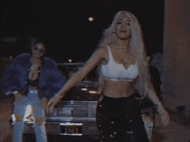Music video gif. Saweetie does a cool, casual dance in front of a shiny black car at night. She's wearing a white crop top and black jeans and another dancer with a purple fur coat is backing her up. 