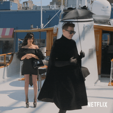 Video gif. A man in a black suit and a long black cape spins around on the set of a photo shoot, holding a tiny black umbrella. A woman in towering high heels and a black feathered dress looks on.