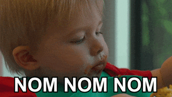 Video gif. Baby with a bib on is eating and begins to feed himself with his left fist but sees his right fist holding a fork coming to his mouth first, so he switches at the last minute and takes a big bite. Text, "Nom nom nom."