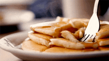 Video gif. Fork dives into a stack of steaming, syrupy pancakes, pulling out a bite.
