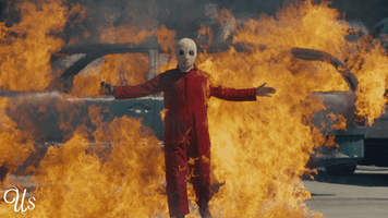 Movie gif. Evan Alex as Pluto in "Us," wearing a red long-sleeved jumpsuit and a white mask, standing in front of a burning car.