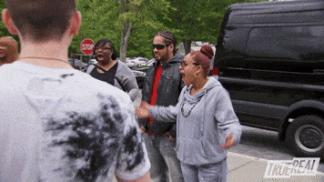 Reality TV gif. We zoom in on a woman in sunglasses with her mouth open in shock and her arms out in a shrug as if to say, "What?"