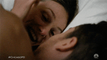 TV gif. Tracy Spiridakos as Hailey and Jesse Lee Soffer as Jay in Chicago PD lay in bed with each other and smile as they kiss. 