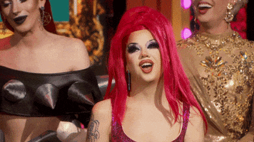 Reality TV gif. Willow Pill, a contestant on RuPaul's Drag Race, has a big red wig on and yellow gloves. She flutters her eyelashes prettily and touches her cheeks to complete the cutesy look.