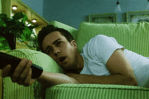 Movie gif. Edward Norton in Fight Club lies on a couch with a TV remote in his hand, looking tired and bored.