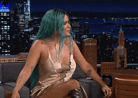 TV gif. Karol G in guest seat on the Tonight Show nods and smiles at the host, saying "yes" and laughing while turning her head to face the audience.