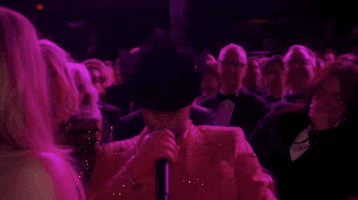 Oscars 2024 GIF. Ryan Gosling begins his performance for I'm Just Ken from Barbie. He sits in the audience and has a black cowboy hat dipped low over his eyes. He takes off the hat and reveals a pair of cat eyed sunglasses and he leans towards Margot Robbie, who sits in front of him. She can't contain her laughter and her glee bubbles up, causing her to cover her mouth with her hand. 