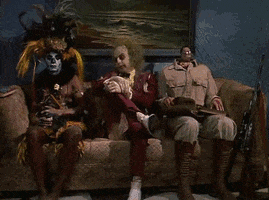 Movie gif. Michael Keaton as Beetlejuice sits between two other ghouls on a couch. He looks down at his number ticket which says, “9, 998, 383, 750, 000.” He looks up at a Now Serving sign that says 2 and then changes to 3. He rolls his eyes and sighs loudly. 