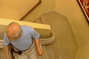 Video gif. Old man slowly descends a staircase on a stair lift.
