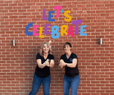 Dvě ženy vyhazující konfety do vzduchu na pozadí zdi s nápisem "Let´s celebrate". 
