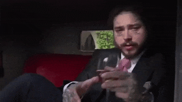 Music video gif. Post Malone in his video for Saint‐Tropez. He's wearing a suit and lounging in a car while raising a glass, toasting us.