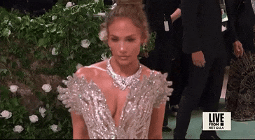 Met Gala 2024 gif. Closeup of Jennifer Lopez in slow motion as she blinks with a calm, neutral expression as cameras flash. She's wearing a near-see-through Schiaparelli sleeveless gown with a plunging v-neckline that culminates in a pattern that flares out from her shoulders like butterfly wings. Her hair is swept up in a high bun with a few wispy strands framing her face.