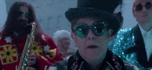 Music video gif. Elton John sings in the music video Merry Christmas with Ed Sheeran swaying in the background with other band members. 