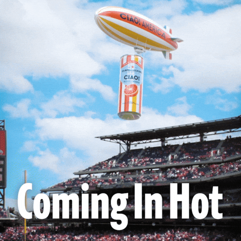 Sponsored gif. Striped blimp carries an enormous striped can of Sanpellegrino Ciao! in the blood orange flavor over a sports stadium filled with people. On the side of the blimp is text that reads, “Ciao! America.” Text below the blimp reads, “Coming in hot.”