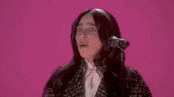 Oscars 2024 GIF. Close up shot of Billie Eilish finishing her performance of "What Was I Made For" on stage at the Oscars. Eilish looks overwhelmed and says, "Whoa!" before laughing and putting a hand up to cover her mouth. 