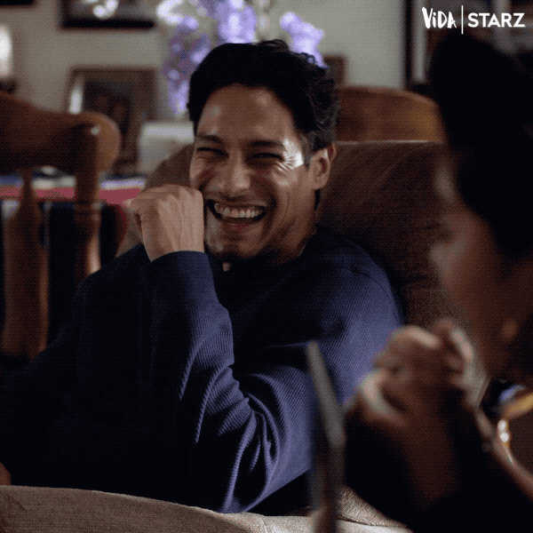 TV gif. Carlos Miranda as Johnny and Chelsea Rendon as Mari in Vida. They're both sitting on the sofa and look at each other before busting up. Mari falls over on the sofa in laughter and Johnny puts his hand up to his mouth, closing his eyes as laughter envelopes them.