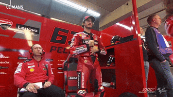 Sports gif. Pecco Bagnaia, a motorcycle racer, waves at us from the garage, decked out in an all red leather racing suit that's covered in logos. An overlay pops up that indicates he's in first grid position. 