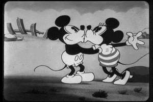 Cartoon gif. Mickey and Minnie are embracing and giving each other sweet kisses. They embrace and cozy up to one another as Minnie flutters her eyelashes at Mickey.