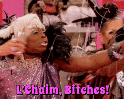 Video gif. Drag queens clinking champagne glasses haphazardly. Text, "L'chaim, bitches!"