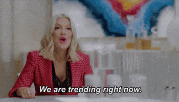 TV gif. Tori Spelling as herself from BH90210 wears a red jacket with a grid pattern. She speaks to us with a smile from across a table. Text, "We are trending right now."
