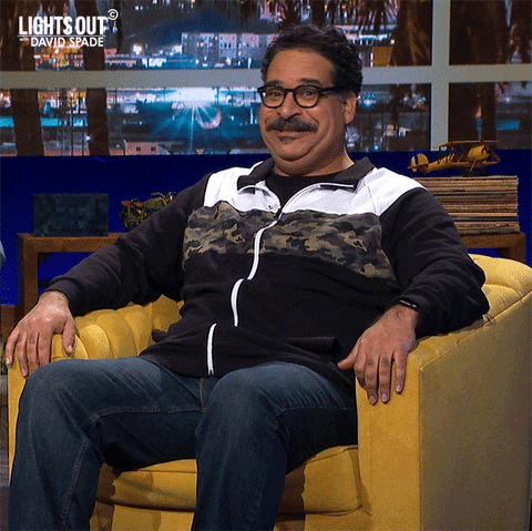 Celebrity gif. Erik Griffin as a guest on Lights Out with David Spade shrugs with a goofy smile.