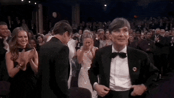 Oscars 2024 GIF. Cillian Murphy walks up to the stage to accept his award for Best Actor while smiling bashfully. Behind him, the crowd gives him a standing ovation. 