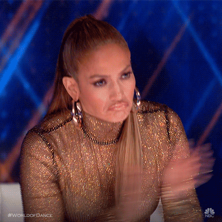 Reality TV gif. Jennifer Lopez as host of World of Dance wears a gold sequin turtleneck and a serious pout as she stands up to clap like she's impressed. 