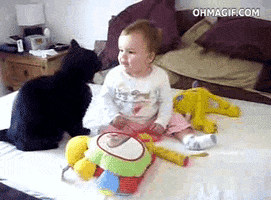 Video gif. Cat paws a baby's nose and the baby laughs, then the cat snuggles up to the baby gently.