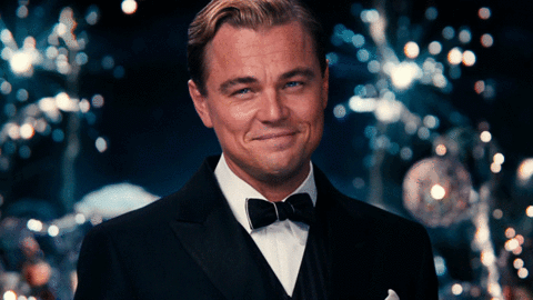 Leonardo DiCaprio, ve filmu The Great Gatsby, zvedající sklenici se šampaňským k přípitku na pozadí vybuchujících ohňostrojů.