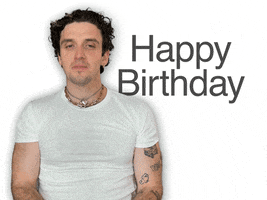 Video gif. Man with tattoos wearing a lot of chain jewelry gestures towards us and says, "Happy birthday" in a sincere yet casual and matter of fact way. 