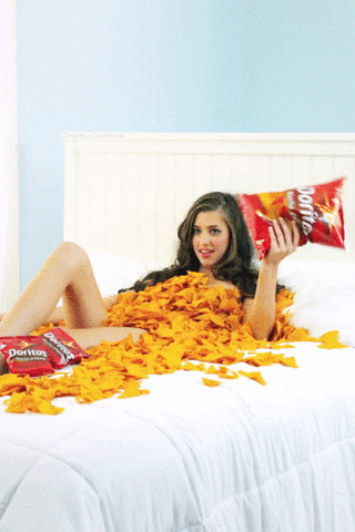 Ad gif. A flirtatious naked woman covered in Doritos lies on a white bed, shaking a bag of Doritos.