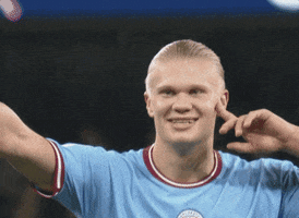 Sports gif. Slow motion video of Erling Haaland wearing a Manchester City jersey as he walks across the field, pointing up at the crowd, smiling and raising a fist in the air. 