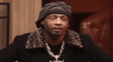 Celebrity gif. Katt Williams wears a gray patterned beanie and a black jacket with a thick chain necklace attached to a silver ship's helm. He leans over and looks at someone bewilderedly before turning away.
