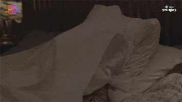 Video gif. A person moves around under the covers on a bed as a person leaps up like a frog in the shadows behind it.
