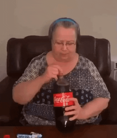 Video gif. An elderly woman places mentos into a large bottle of coke. She panics as the soda spews all over her.