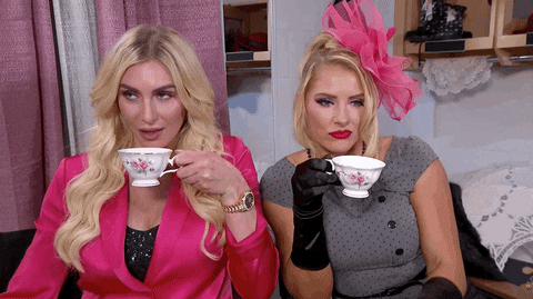 Celebrity gif. Wrestlers Lacey Evans and Charlotte Flair wear fancy clothes while sipping tea from white cups. They look around with alluring eyes like they're ready for some juicy gossip.