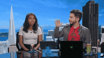 Celebrity gif. Sitting at a round table hosting their show, Mica Burton and Andy Cortez high five each other, from which Mica recoils in pain, examining her hand.