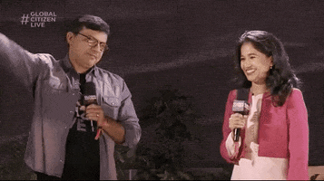 TV gif. Man and woman hosts on Global Citizen Live wave and smile while holding their microphones.