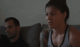 Sports gif. Mayra Bueno Silva sits on a couch next to a man while looking forward nervously and clapping awkwardly.