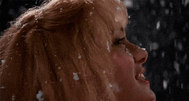 Movie gif. Winona Ryder as Kim in Edward Scissorhands smiles while looking up and turning around slowly as snow falls.