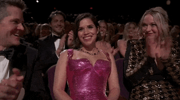 Oscars 2024 GIF. America Ferrera, seated at the Oscars, girlishly accepts praise, flanked by people applauding and looking at her with pride.