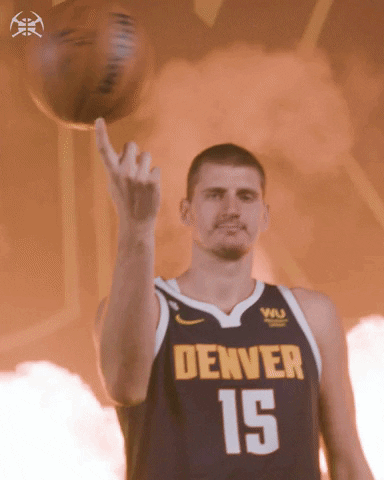 Sports gif. Nikola Jokic of the Denver Nuggets spins a basketball on one hand, looks at us with a vacant expression as flames burst in the background. 