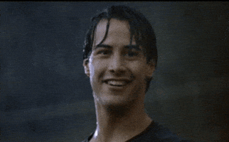 Movie gif. Keanu Reeves as Johnny Utah in Point Break smiles and gives a cool thumbs-up while standing soaking wet from the pouring rain around him.