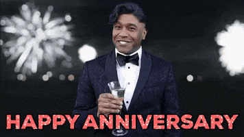 Meme gif. A man holds a glass full of champagne, motion to us with a "cheers" motion, in front of a background of bursting fireworks, in the style of the Leo DiCaprio "Great Gatsby" meme. The words "Happy Anniversary" appear as text.